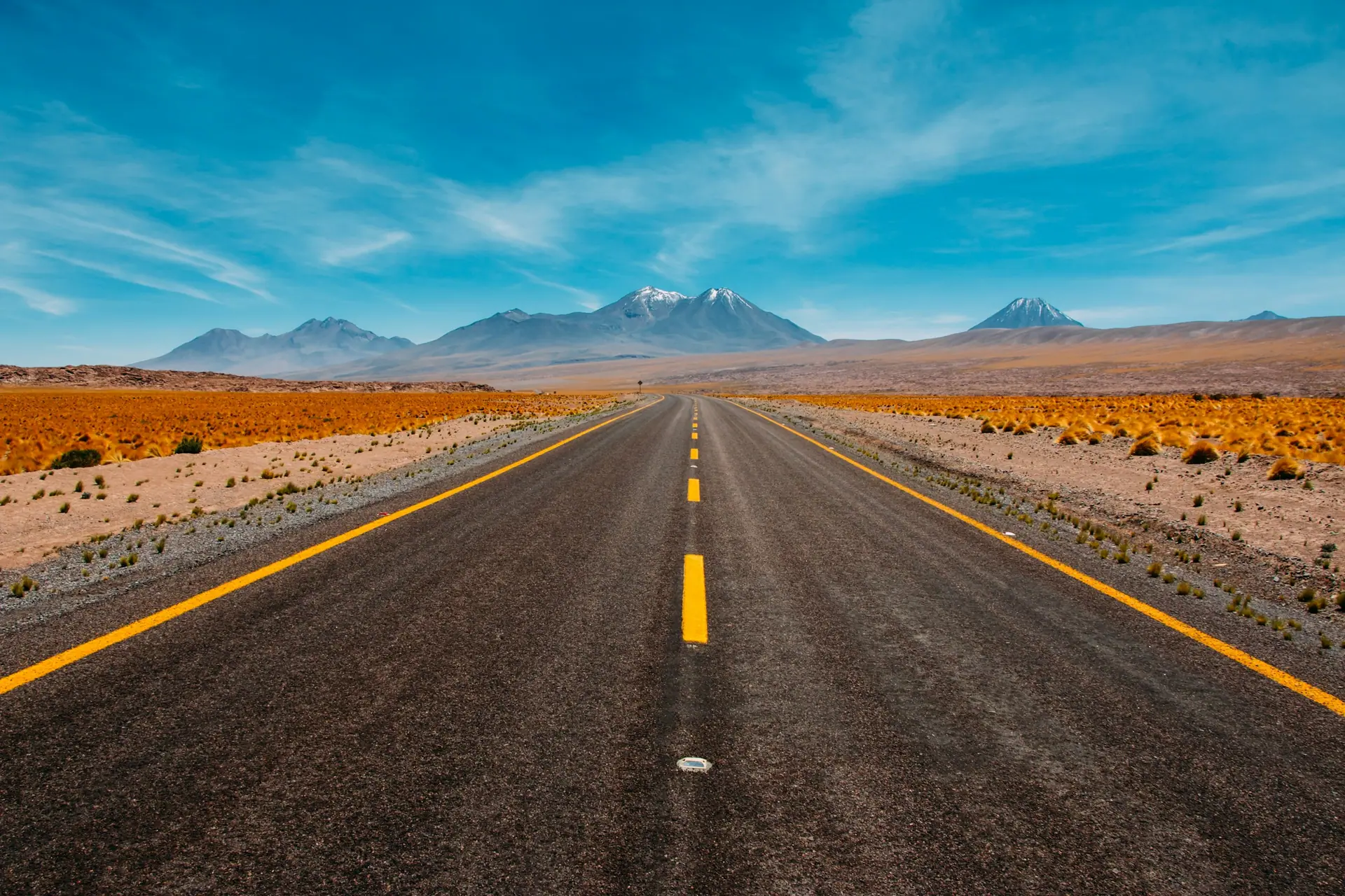 Empty sealed road with yellow line markings running through desert towards snow-capped mountains